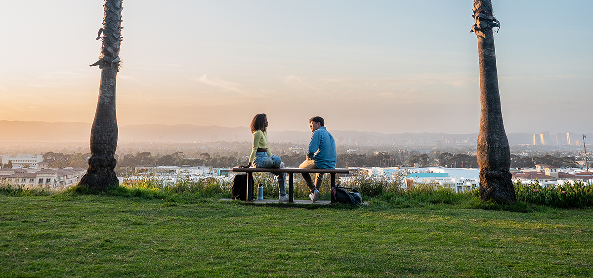 Two students sitting on a bench on a cliff overlooking Playa Vista with a sunset behind them.
